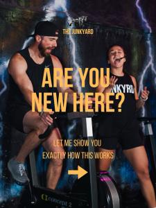 Two people cycling in a Junk Yard Fitness class with a promotional graphic reading “Are you new here? Let me show you exactly how this works.”