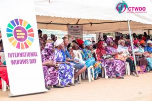 A photo of a marquee in Kenya, with people sitting down, and a sign in the corner saying 'International Widows Day'