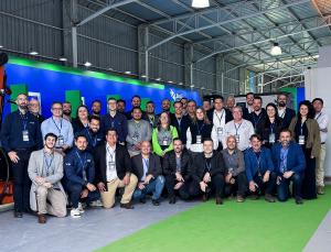 Un grupo de profesionales de la delegación brasileña de Brazil Machinery Solutions posa para una foto colectiva en el interior de una feria internacional, con el stand al fondo en tonos azul y verde, estructura de pabellón industrial e iluminación suspend