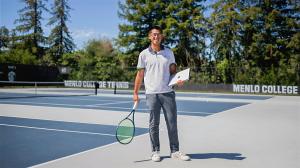 A student stands on an outdoor tennis court holding a tennis racket and a laptop, with Menlo College tennis courts and trees visible in the background.
