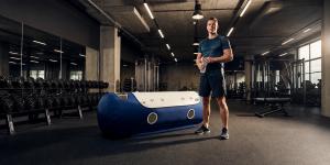 Athlete standing beside a hyperbaric oxygen chamber in a modern gym, showcasing advanced recovery technology for sports performance and wellness.
