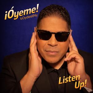 A man in a black suit has his two hands held up to his ears to better capture the sounds surrounding him. He is pictured against a blue-purple background and the album title Listen Up! can be seen in the bottom right corner of the album cover