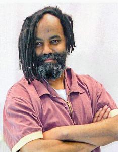 Mumia stands with his arms folded and a strong smile