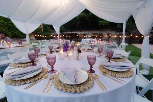 Outdoor wedding reception table styled by Wedding Revolution LLC with white linens, gold flatware, woven chargers, purple goblets, and floral centerpieces under a draped canopy in Orange County California