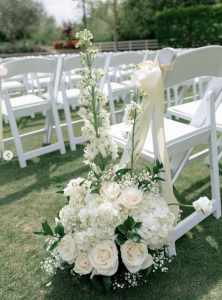 Outdoor wedding ceremony with white folding chairs and a large white floral arrangement of roses hydrangeas and baby's breath along the aisle styled by Wedding Revolution LLC in Orange County California