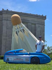 Aaron Novinger stands next to his blue velomobile, known as America's weirdest bike, in front of a giant shuttlecock sculpture in Kansas City during the Pedaling Against Ponzis charity ride.