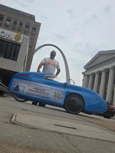 Aaron Novinger stands behind his blue velomobile, a fully enclosed bicycle, on a city street with the iconic St. Louis Gateway Arch in the background during the Pedaling Against Ponzis charity ride.