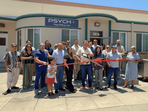 Smiling group of people of all ages standing in front of PsychPlus clinic performing ribbon cutting ceremony.