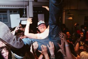 An employee from Nudie Jeans crowd surfing during the first installment of Tomorrow's Tracks in Soho, London. He is wearing Nudie jeans, a white t-shirt and a hat while being lifted by the crowd and he has jeans behind and smile towards the camera.