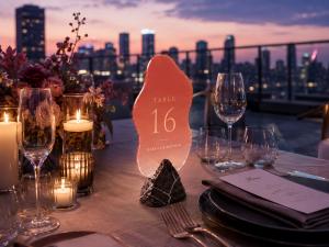 A close-up photo of a luxury outdoor evening event table setup. The centerpiece is a wave-shaped translucent dyed acrylic table number sign with a soft pink gradient transition, mounted on a dark marble base, with the text "TABLE 16" and "ISABELLE & MATTH