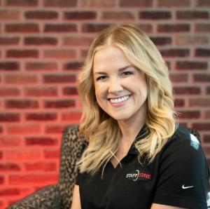 Corporate portrait of Amanda Albert smiling in a black Staff Zone polo shirt in front of a red brick wall