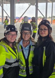 Three women wearing hard hats and high-visibility safety vests smiling together at a DFW construction site during Safety Stand Down week, standing beneath exposed steel beams with active jobsite work visible in the background.