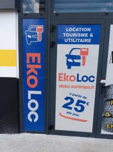 Enseigne et porte d'entrée de l'agence EkoLoc Martinique, spécialisée dans la location de véhicules de tourisme et utilitaires. La vitrine affiche les tarifs compétitifs de l'agence, à partir de 25 € par jour, et son site internet ekoloc-martinique.fr.