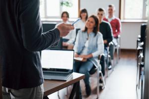 Un formateur s'adresse à un groupe de participants assis en classe, lors d'une séance de formation en entreprise. Au premier plan, un ordinateur portable ouvert sur une table; en arrière-plan, les participants souriants écoutent attentivement, illustrant