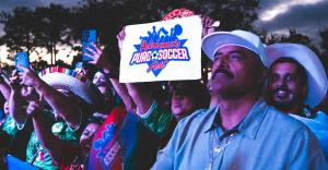 A crowd of fans wearing Mexico soccer jerseys and traditional hats cheers while holding up an Adriana's Puro Soccer Fest sign during the soccer watch party in Downtown Chula Vista