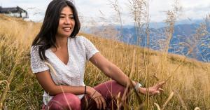A woman with long dark hair sits in a field of tall golden grass on a hillside, smiling gently with mountains visible in the distance.
