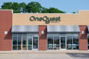 The exterior of the OneQuest Health outpatient facility in Grant County. The building has a modern facade combining red and tan brickwork. A dimensional green sign reading "OneQuest Health" is mounted above wide glass windows.