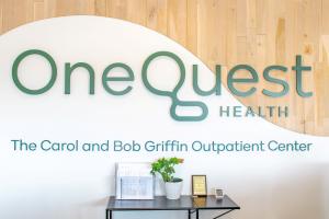 A bright, modern office lobby interior featuring a prominent dimensional sign on a curved white wall that transitions into light wood paneling. The green text reads "OneQuest Health" above the phrase "The Carol and Bob Griffin Outpatient Center."