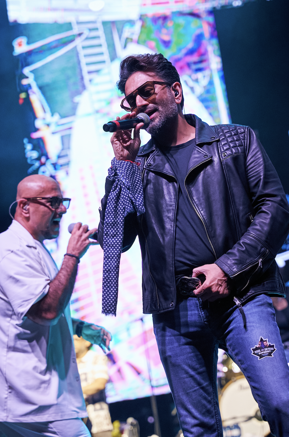 Vishal & Sheykhar performing on stage with microphones, colorful LED visuals, and live concert production for “The Superhit Tour,” supported in select markets by Kash Patel Productions.