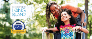 A smiling caregiver stands behind a child using a wheelchair outdoors, alongside the Discover Long Island logo and the Certified Autism Center™ badge.