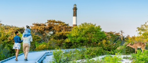 Family walking on a boardwalk near green trees and a lighthouse, with a deer standing on the right side in a natural setting.