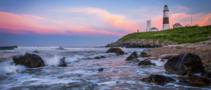 Seaside view of a lighthouse on a rocky shore at sunset with waves crashing and a colorful sky.