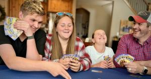 Four members of a family laugh around a kitchen table while playing the Skip the Salad card game, each holding a fanned hand of brightly colored Skip the Salad cards.
