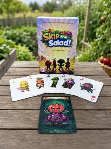 The Skip the Salad card game box stands on a wooden table in a garden, with five illustrated vegetable character cards fanned in front and a Rotten Tomato card below.