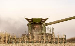 A large, green John Deere combine harvester moving forward through a dry cornfield during harvest season. An operator is visible inside the glass cab. A massive corn head attachment at the front cuts through rows of stalks, kicking up a thick cloud of dus