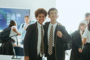 Two smiling teenage students in The Independent School of Jakarta senior uniform, black blazers and striped ties, in a modern classroom with a smartboard and classmates behind them.