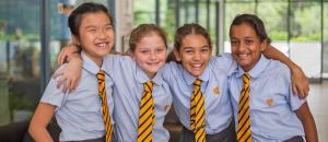 Four smiling primary-age pupils in The Independent School of Jakarta uniform, blue shirts and gold-and-navy striped ties with the ISJ lion crest, arms around each other.
