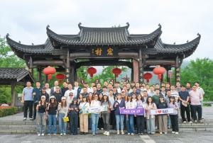 A large group of about 60 DAS employees stands in front of a traditional Chinese gate with curved roofs and red lanterns. The group is standing on a flight of stairs and holding several banners, including “DAS CN 25” and “I ❤️ DAS.” Green trees and a clou