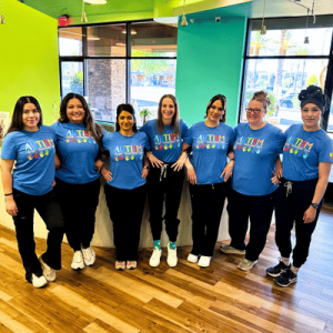 Group of seven women standing indoors, wearing matching blue "Autism Awareness" t-shirts with colorful handprints, smiling at the camera.