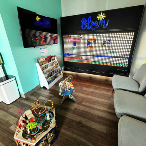 Waiting area with child activity toys, seating, a large Lite-Brite wall art, and a magazine rack under a mounted TV.