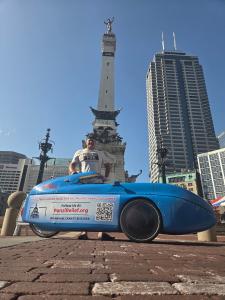 Aaron Novinger stands with his bright blue velomobile in front of the Soldiers and Sailors Monument in downtown Indianapolis while on his 2,000-mile ride supporting Ponzi scheme victims.
