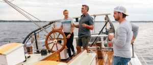 Three men on a sailboat, one steering the wheel, another talking, and the third holding a cup, with water and shoreline in the background.