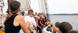 Group of people working together to hoist sails on a sailboat in calm waters on a clear day.