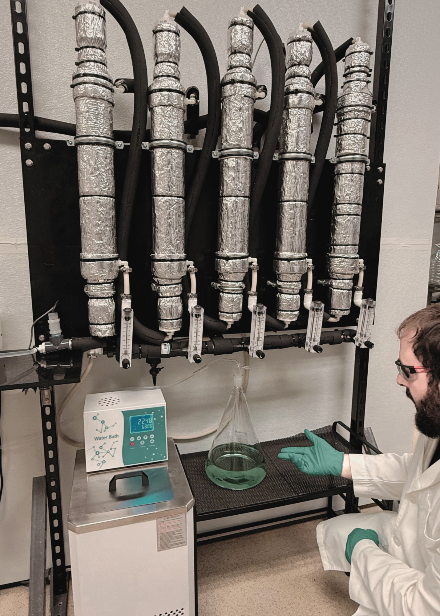 Brandon Carpenter in a lab coat and safety glasses, adjusting equipment beside five foil-wrapped refining columns, with a flask of green nickel sulfate solution on a stand below.
