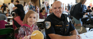 A Redmond police officer sits beside a smiling child at a community event, with attendees gathered in the background.