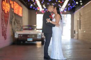 A newly married couple in tux and gown kiss in a drive thru wedding venue in Las Vegas with a white convertible vintage Cadillac in the background