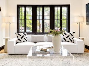 Bright historic living room professionally staged with a white modern sofa, acrylic coffee table, black-framed windows, and neutral décor by Jsquared Interior Staging & Design.