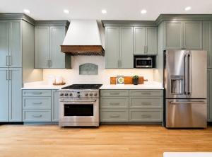Luxury kitchen featuring soft green shaker cabinets, a custom wood vent hood, white countertops, stainless steel appliances, and professional home staging by Jsquared Interior Staging & Design.