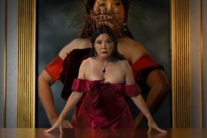 An older Asian woman looks directly into the camera. She is leaning her hands on a table. She is dressed in a silky red off shoulder dress with black pants. She is wearing a necklace and earrings and has long dark hair. Behind her in her golden palace wit