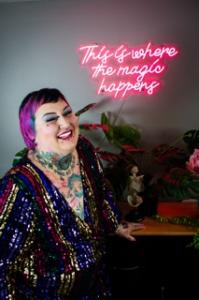 A close-up portrait of Wednesdae Reim Ifrach smiling brightly, showcasing short magenta and teal hair, intricate neck and chest tattoos, and a dazzling, multi-colored sequined jacket