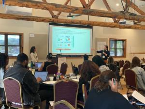 Participants of the Community Builders program convene in a conference room. Two people at the front of the room share a slide presentation, while others at small tables watch and take notes.