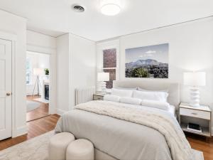Modern primary bedroom with neutral upholstered bed, layered white bedding, designer lamps, contemporary landscape artwork, and hardwood floors.