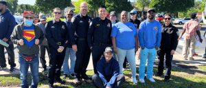 Group of people, including police officers and civilians, smiling outdoors on a sunny day in a community setting.