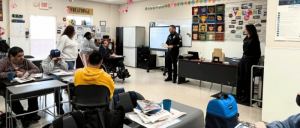 Classroom setting with a police officer speaking to seated students, a woman standing nearby, and educational materials on walls and tables.