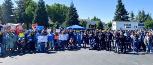 Large diverse group of people gathered outdoors holding signs, with trees and a blue canopy tent in the background on a sunny day.