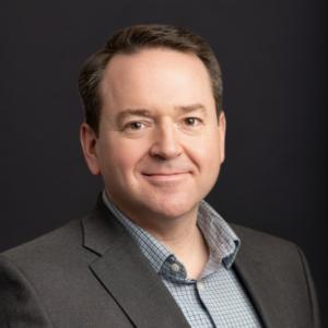 Headshot of Brad Sprague, Director of Sales & Marketing at ProInfoNet, smiling and wearing a suit jacket in front of a grey background.